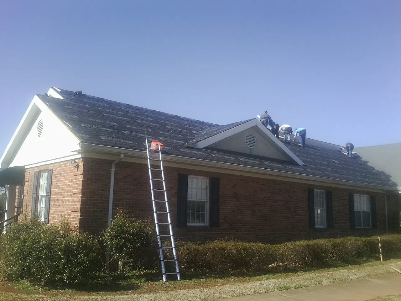 Crew tearing shingles off a brick ranch roof with a ladder set — Emergency Roof Repair work by our crew in Rowan
