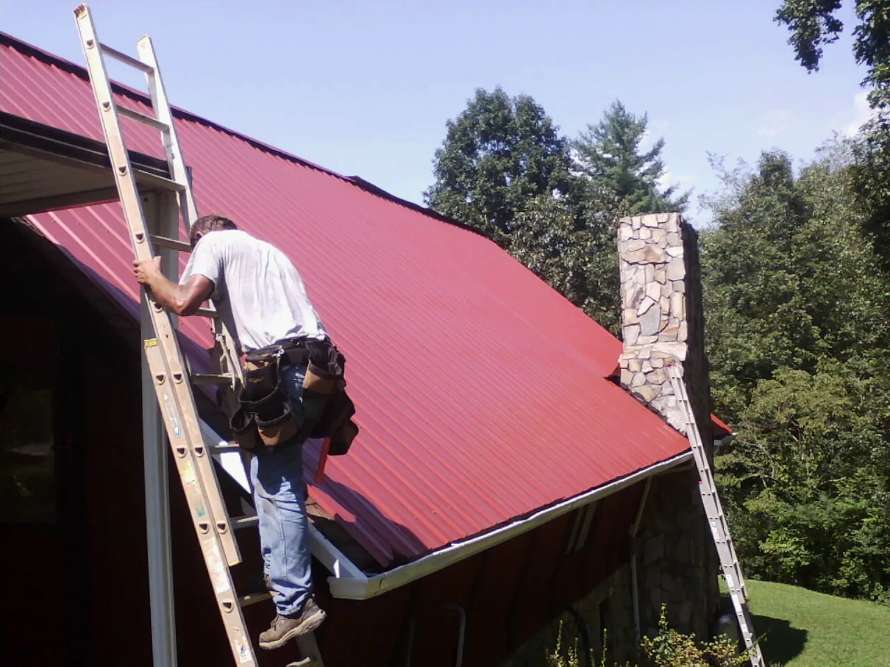 Roofer on a ladder beside a red metal roof and stone chimney — Soffit & Fascia Repair by Roofing Brothers in Rowan